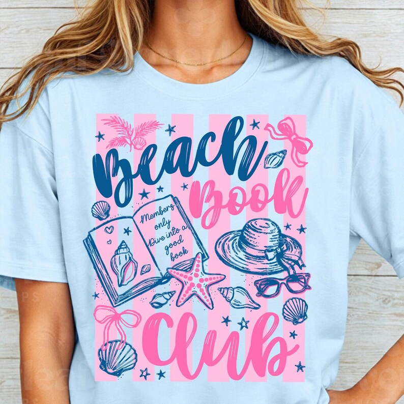 May include: Light blue t-shirt with a pink and white striped design. The shirt features the words "Beach Book Club" in a script font, along with illustrations of seashells, a starfish, a hat, and sunglasses. The text "Members only dive into a good book" is also included.