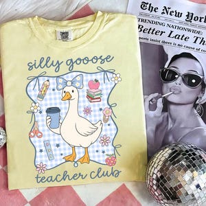 May include: A light yellow Comfort Colors t-shirt with a cartoon goose design. The goose holds a coffee cup and is surrounded by school-themed graphics and the words "silly goose teacher club". A newspaper and disco ball are in the background.