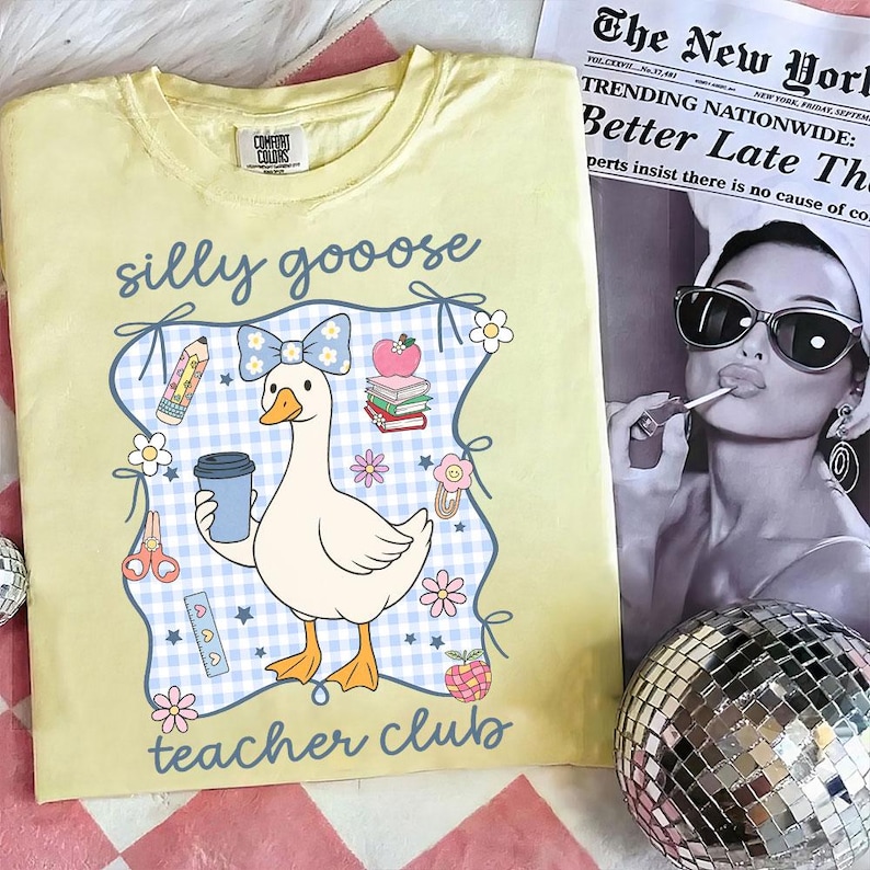 May include: A light yellow Comfort Colors t-shirt with a cartoon goose design. The goose holds a coffee cup and is surrounded by school-themed graphics and the words "silly goose teacher club". A newspaper and disco ball are in the background.