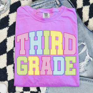 May include: A bright pink short-sleeved t-shirt with a crew neckline and a Comfort Colors brand label. The front features the text THIRD GRADE in large, block-style letters with a vertical white striped pattern in various colors including blue, yellow, and green.