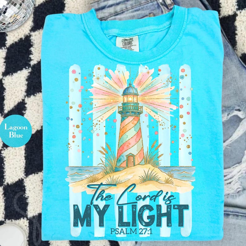 Pu&ograve; includere: T-shirt blu laguna con un'illustrazione colorata di un faro. Il faro ha un design a strisce ed &egrave; ambientato su un'isola sabbiosa. Il testo "The Lord is MY LIGHT" e "PSALM 27:1" &egrave; stampato sotto.