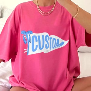 May include: A bright pink t-shirt featuring a white pennant with the word "CUSTOM" in light blue. A light blue bow is tied to the pennant. The shirt has short sleeves and a relaxed fit.