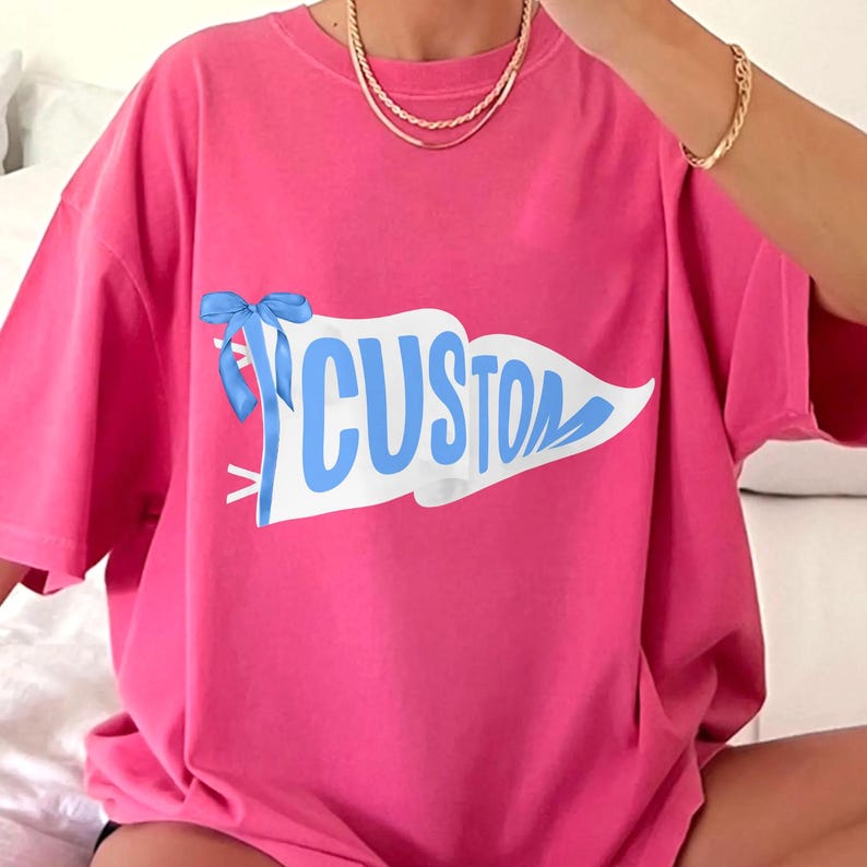 May include: A bright pink t-shirt featuring a white pennant with the word "CUSTOM" in light blue. A light blue bow is tied to the pennant. The shirt has short sleeves and a relaxed fit.