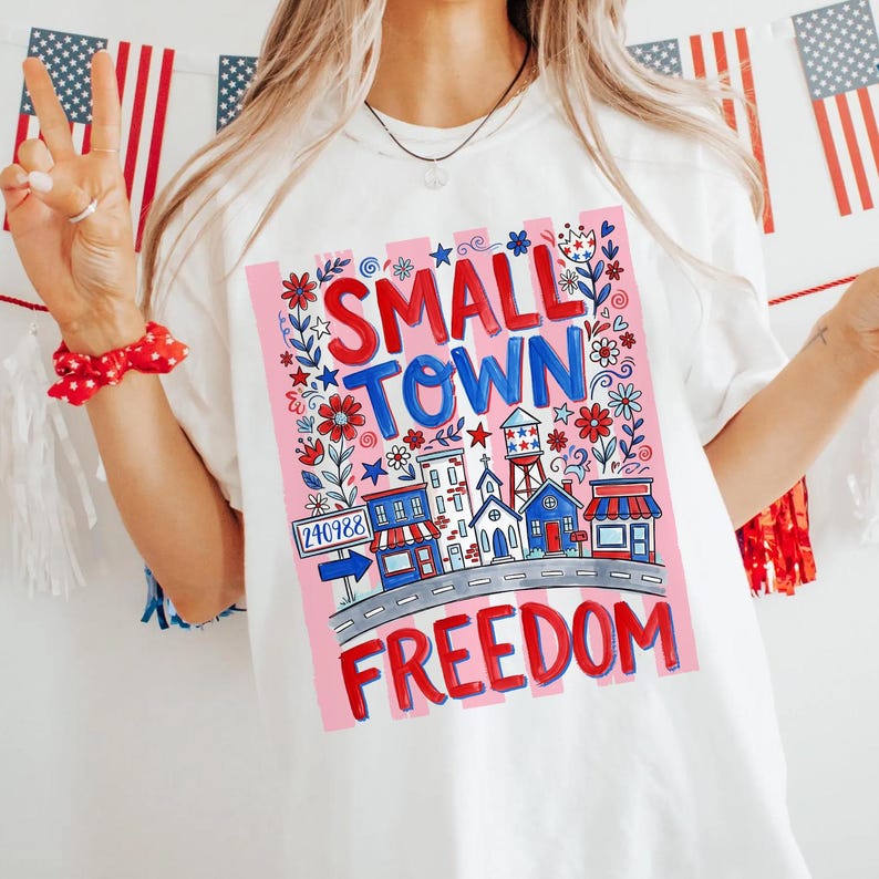 May include: White t-shirt with a colorful graphic design. The design features the words "SMALL TOWN FREEDOM" in red and blue, surrounded by floral and architectural illustrations. The shirt has a crew neck.