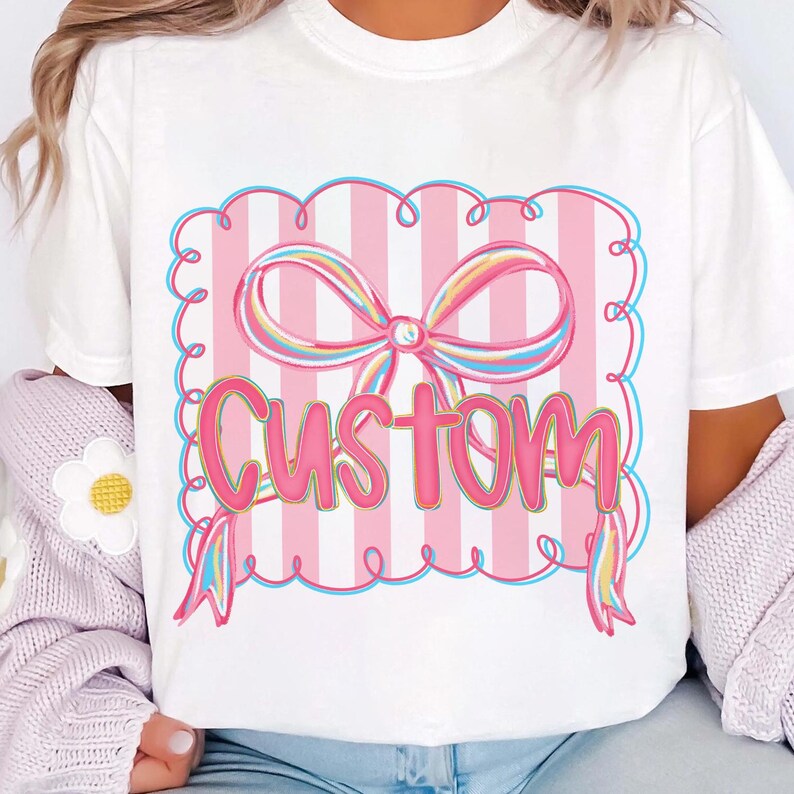 May include: White t-shirt with a pink bow and the word "Custom". The design features pink and white stripes, a blue outline, and rainbow-coloured accents on the bow. A fun and colourful design.