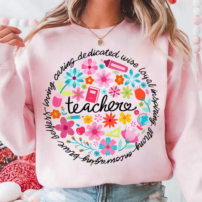 May include: Pink sweatshirt with a circle design featuring the word "teacher" surrounded by colorful flowers, leaves, and school supplies. The text around the circle reads "caring, dedicated, wise, loyal, inspiring, strong, brave, loving".