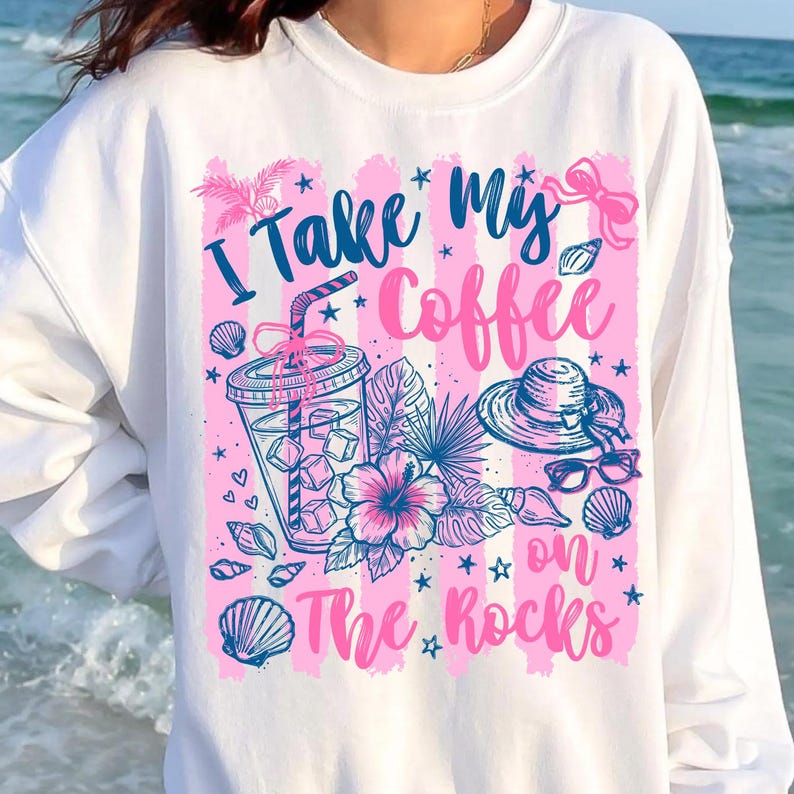 May include: White sweatshirt with a pink and blue graphic. The graphic includes the text "I Take My Coffee on The Rocks" along with illustrations of a drink, hat, sunglasses, seashells, and tropical flowers.