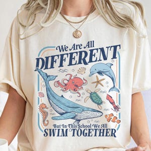 May include: Cream-colored t-shirt with a graphic featuring various sea creatures, including a whale, octopus, and dolphin. The text reads "We Are All DIFFERENT But In This School We All SWIM TOGETHER."