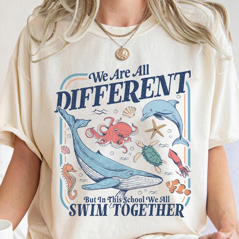 May include: Cream-colored t-shirt with a graphic featuring various sea creatures, including a whale, octopus, and dolphin. The text reads "We Are All DIFFERENT But In This School We All SWIM TOGETHER."