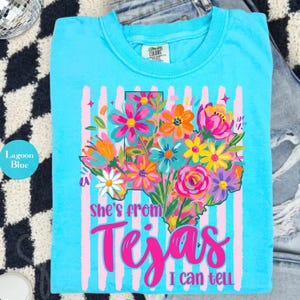 May include: Lagoon blue t-shirt with a floral Texas graphic. The design features a colorful bouquet of flowers in the shape of the state, with the text "She's from Tejas I can tell" in bright pink.