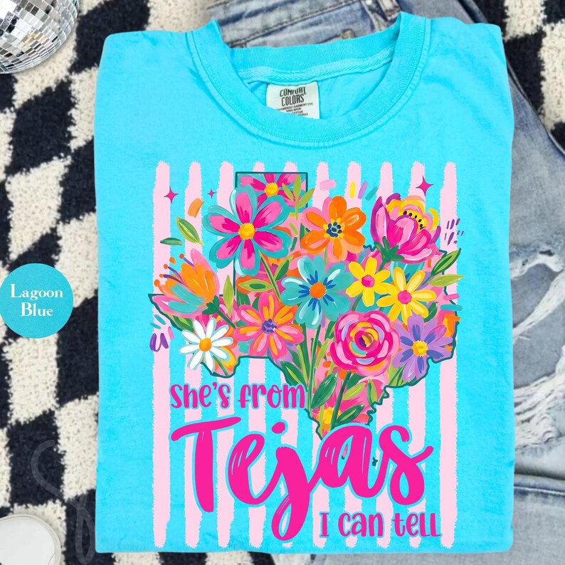 May include: Lagoon blue t-shirt with a floral Texas graphic. The design features a colorful bouquet of flowers in the shape of the state, with the text "She's from Tejas I can tell" in bright pink.