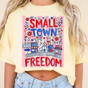 May include: A light yellow t-shirt featuring a colorful graphic design. The design includes the words "SMALL TOWN FREEDOM" in red and blue, along with illustrations of buildings, flowers, and stars. A road sign with the number "240988" is also included.