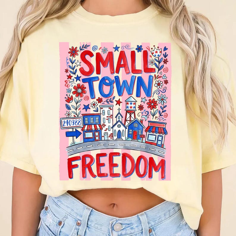 May include: A light yellow t-shirt featuring a colorful graphic design. The design includes the words "SMALL TOWN FREEDOM" in red and blue, along with illustrations of buildings, flowers, and stars. A road sign with the number "240988" is also included.
