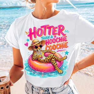 K&ouml;nnte beinhalten: Wei&szlig;es T-Shirt mit einem Grafikdesign eines Geparden mit Sonnenbrille und Strohhut, der auf einem rosa Schwimmring im Wasser liegt. Der Text "HOTTER THAN A HOOCHIE COOCHIE" steht in Pink und Orange &uuml;ber dem Geparden.