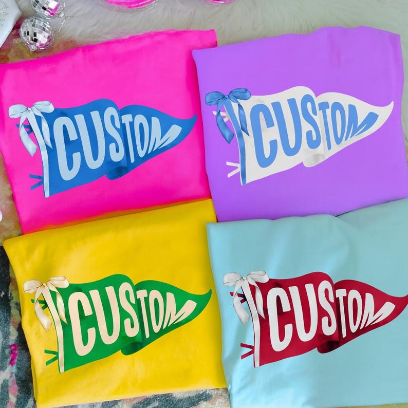 May include: Four folded t-shirts in bright colors: pink, purple, yellow, and light blue. Each shirt features a pennant design with the word "CUSTOM" in a contrasting color. The pennants are adorned with bows.