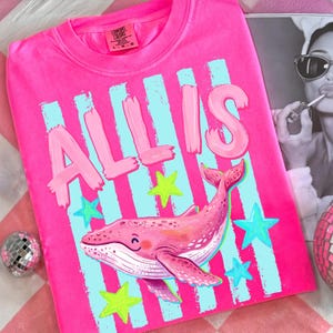 以下が含まれることがあります： 明るいピンク色のTシャツ。名前「ALLIS」は、薄いピンク色の筆記体で書かれています。前面には、笑顔の顔と青と緑の星のデザインが施されたピンク色のクジラが描かれています。このシャツはComfort Colorsブランドです。