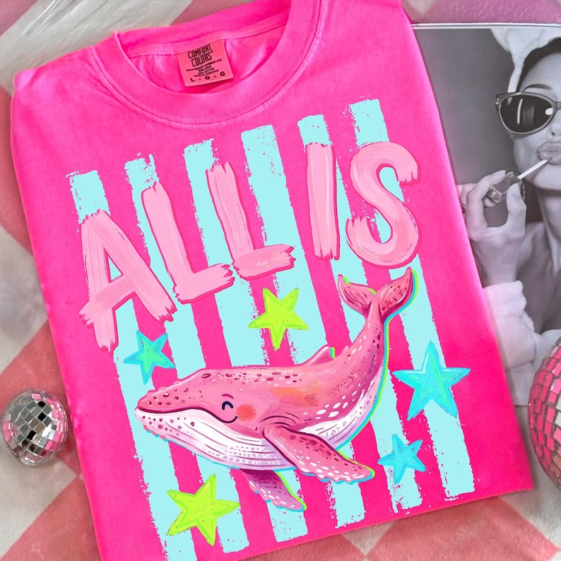 以下が含まれることがあります： 明るいピンク色のTシャツ。名前「ALLIS」は、薄いピンク色の筆記体で書かれています。前面には、笑顔の顔と青と緑の星のデザインが施されたピンク色のクジラが描かれています。このシャツはComfort Colorsブランドです。