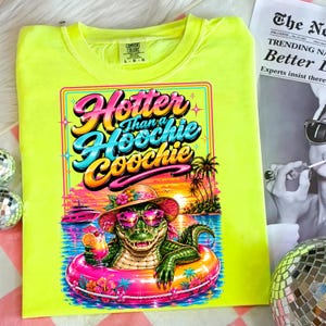 May include: Neon yellow Comfort Colors t-shirt with a graphic of an alligator in sunglasses and a hat, floating in a pink inner tube. The graphic reads "Hotter Than a Hoochie Coochie" in colorful text. A tropical sunset scene is in the background.
