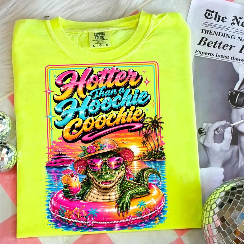May include: Neon yellow Comfort Colors t-shirt with a graphic of an alligator in sunglasses and a hat, floating in a pink inner tube. The graphic reads "Hotter Than a Hoochie Coochie" in colorful text. A tropical sunset scene is in the background.
