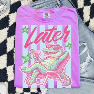 Pode incluir: Uma camiseta lil&aacute;s Comfort Colors com um gr&aacute;fico de um jacar&eacute; de &oacute;culos de sol relaxando em uma cadeira de praia, segurando um coquetel. A palavra "Later" est&aacute; escrita em rosa acima do jacar&eacute;, com estrelas verdes.