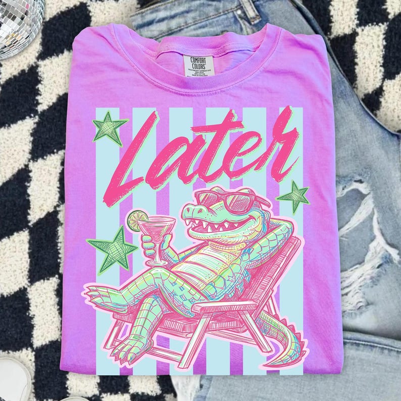 Pode incluir: Uma camiseta lil&aacute;s Comfort Colors com um gr&aacute;fico de um jacar&eacute; de &oacute;culos de sol relaxando em uma cadeira de praia, segurando um coquetel. A palavra "Later" est&aacute; escrita em rosa acima do jacar&eacute;, com estrelas verdes.