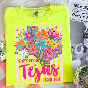 May include: Neon yellow t-shirt with a floral Texas outline design. The design features a colorful bouquet of flowers in shades of pink, blue, orange, and white. Text on the shirt reads "She's from Tejas I can tell."