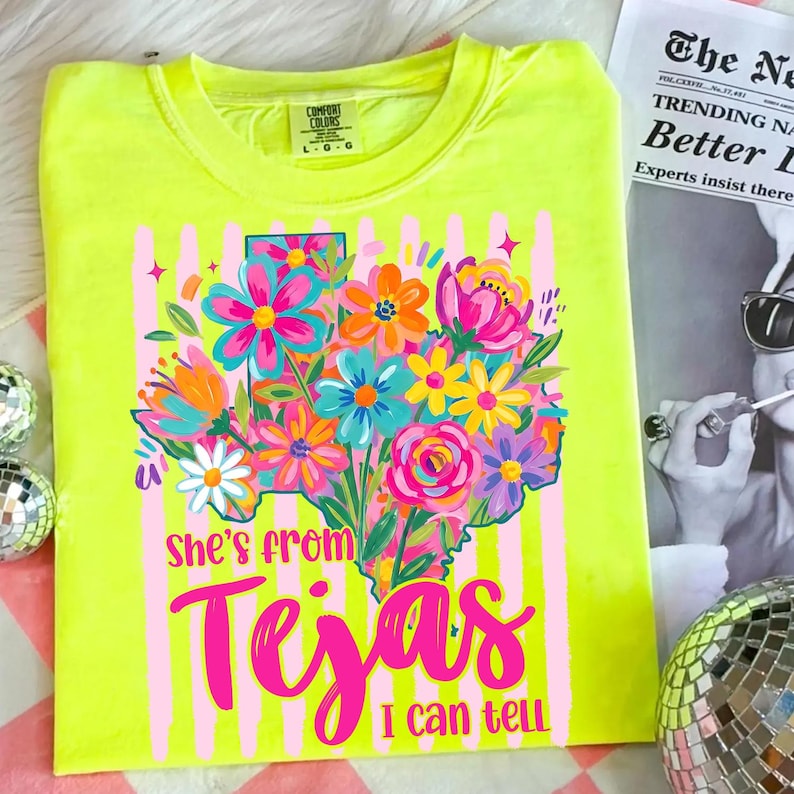 May include: Neon yellow t-shirt with a floral Texas outline design. The design features a colorful bouquet of flowers in shades of pink, blue, orange, and white. Text on the shirt reads "She's from Tejas I can tell."