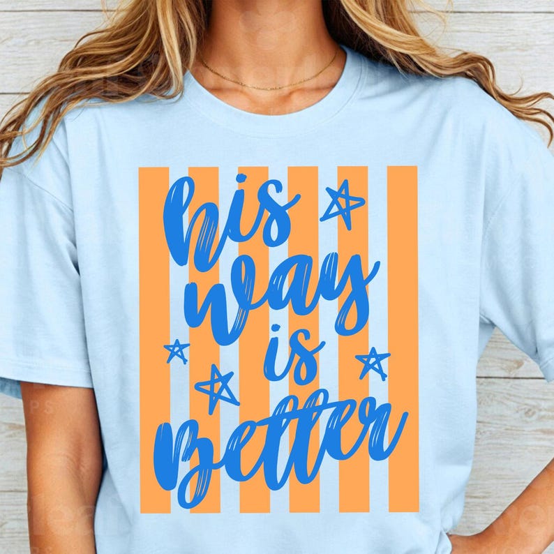 May include: Light blue t-shirt with the text "His Way Is Better" in blue script. The text is set against a background of orange vertical stripes. The shirt is a casual wear item.