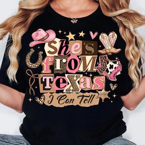 May include: Black t-shirt with a graphic design that reads "She's From Texas I Can Tell." The design includes a pink cowboy hat, boots, a horseshoe, and the outline of the state of Texas.