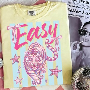 Pode incluir: Camiseta amarela Comfort Colors com um gr&aacute;fico rosa e azul. O design apresenta a palavra "EASY" com la&ccedil;os, um tigre, estrelas e listras azuis. Uma pe&ccedil;a de roupa casual.