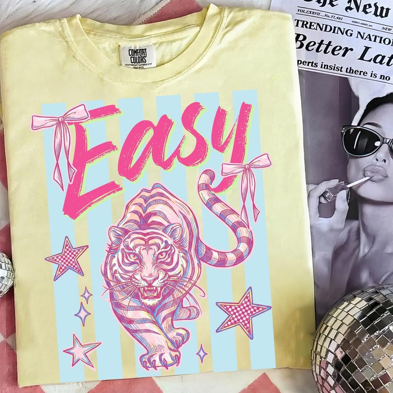 Pode incluir: Camiseta amarela Comfort Colors com um gr&aacute;fico rosa e azul. O design apresenta a palavra "EASY" com la&ccedil;os, um tigre, estrelas e listras azuis. Uma pe&ccedil;a de roupa casual.