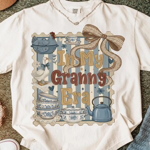 以下が含まれることがあります： ヴィンテージスタイルのグラフィックが施された白いTシャツ。デザインは、ガチョウ、鶏、ティーカップ、ボウルのイラストが描かれた青とベージュの切手です。「In My Granny Era」と書かれています。
