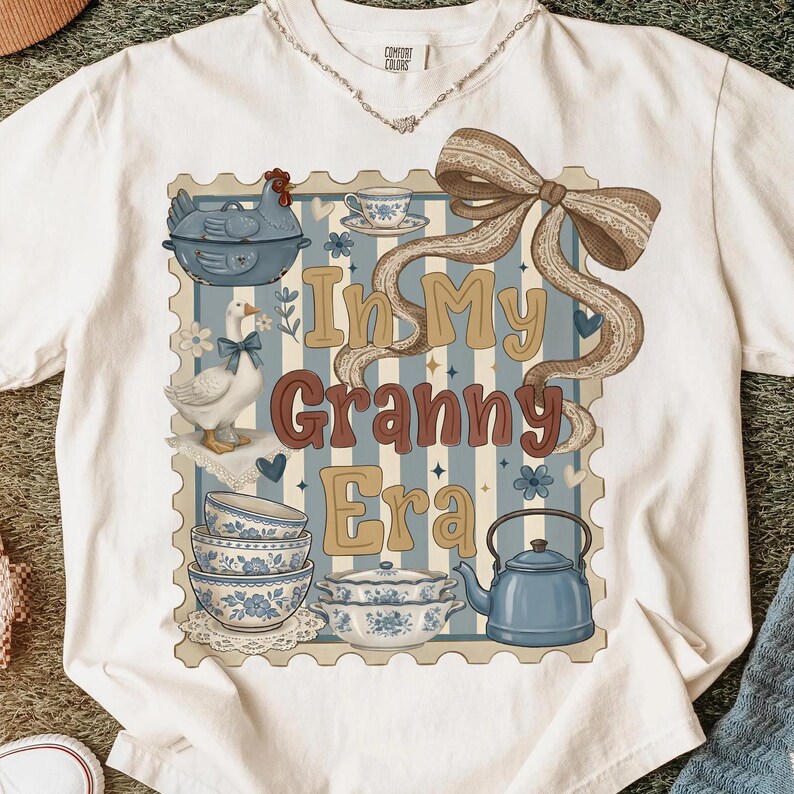 以下が含まれることがあります： ヴィンテージスタイルのグラフィックが施された白いTシャツ。デザインは、ガチョウ、鶏、ティーカップ、ボウルのイラストが描かれた青とベージュの切手です。「In My Granny Era」と書かれています。