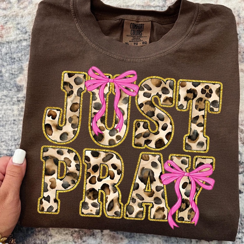 May include: Brown t-shirt with the words "JUST PRAY" in a leopard print design outlined in gold. Pink bows are placed above the letters "U" and "R" and below the letter "Y". The shirt is folded.