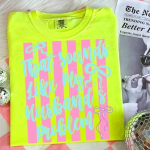 May include: Neon yellow t-shirt with pink vertical stripes and the phrase "That sounds like my husband's problem" in light blue script. The shirt has a crew neck and short sleeves. The Comfort Colors brand label is visible.