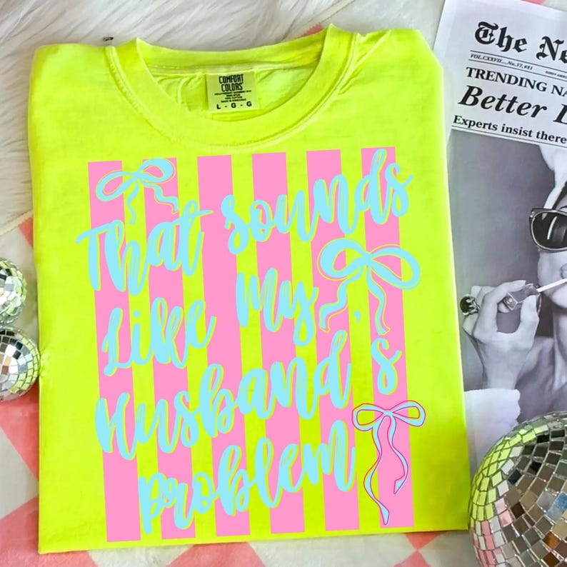 May include: Neon yellow t-shirt with pink vertical stripes and the phrase "That sounds like my husband's problem" in light blue script. The shirt has a crew neck and short sleeves. The Comfort Colors brand label is visible.