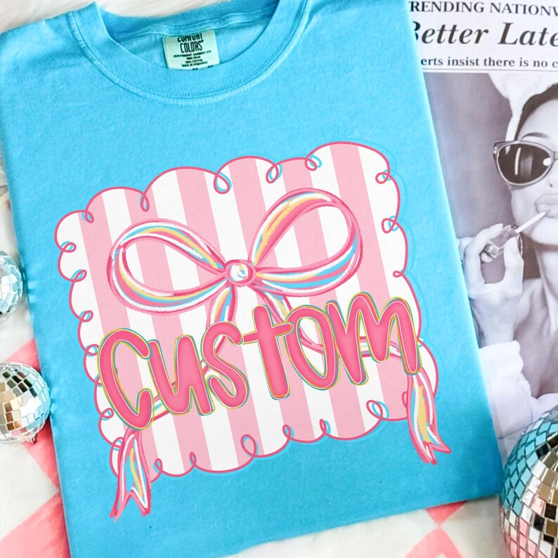 May include: A pale blue t-shirt with a pink and white striped design featuring a bow and the word "CUSTOM" in pink. The bow has rainbow-coloured accents. The shirt has a Comfort Colors tag.