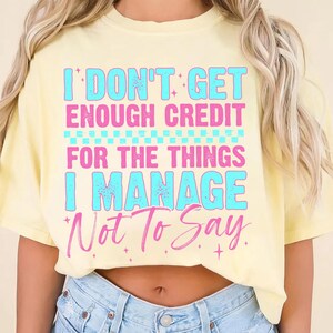 May include: A light yellow t-shirt with the text "I DON'T GET ENOUGH CREDIT FOR THE THINGS I MANAGE Not To Say" in blue and pink. The shirt is a casual style.