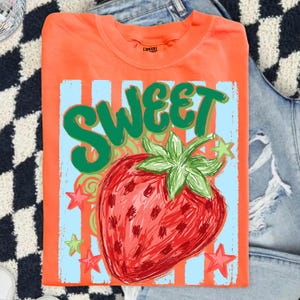 May include: Orange t-shirt with a large strawberry graphic and the word "SWEET" in green. The strawberry is red with green leaves, set against a light blue background with orange stripes and stars. The shirt is folded.