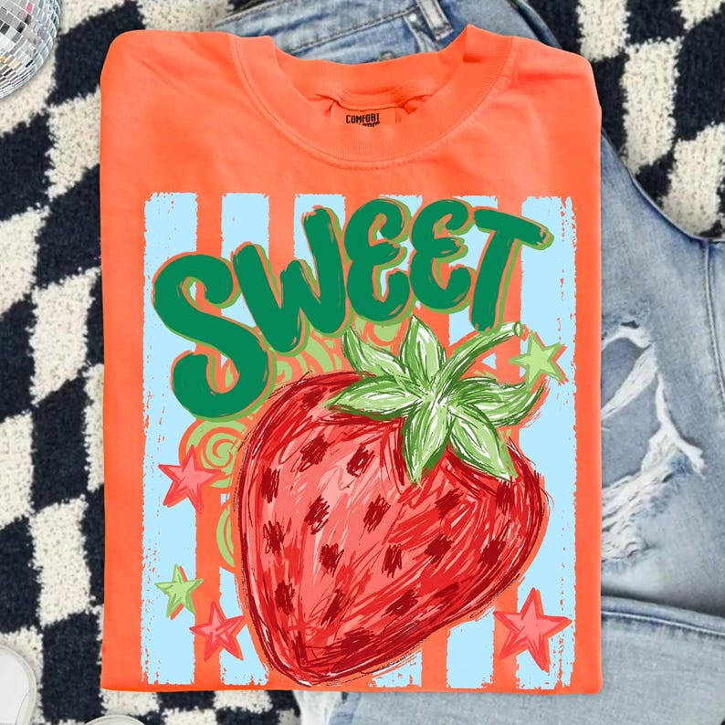 May include: Orange t-shirt with a large strawberry graphic and the word "SWEET" in green. The strawberry is red with green leaves, set against a light blue background with orange stripes and stars. The shirt is folded.
