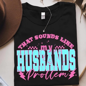 May include: Black t-shirt with the text "THAT SOUNDS LIKE my HUSBANDS Problem" in a distressed, retro font. The text is in shades of blue and pink. A silver necklace with a small pendant is visible.
