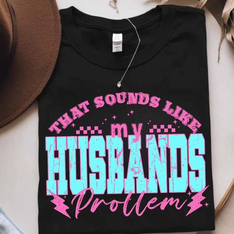 May include: Black t-shirt with the text "THAT SOUNDS LIKE my HUSBANDS Problem" in a distressed, retro font. The text is in shades of blue and pink. A silver necklace with a small pendant is visible.
