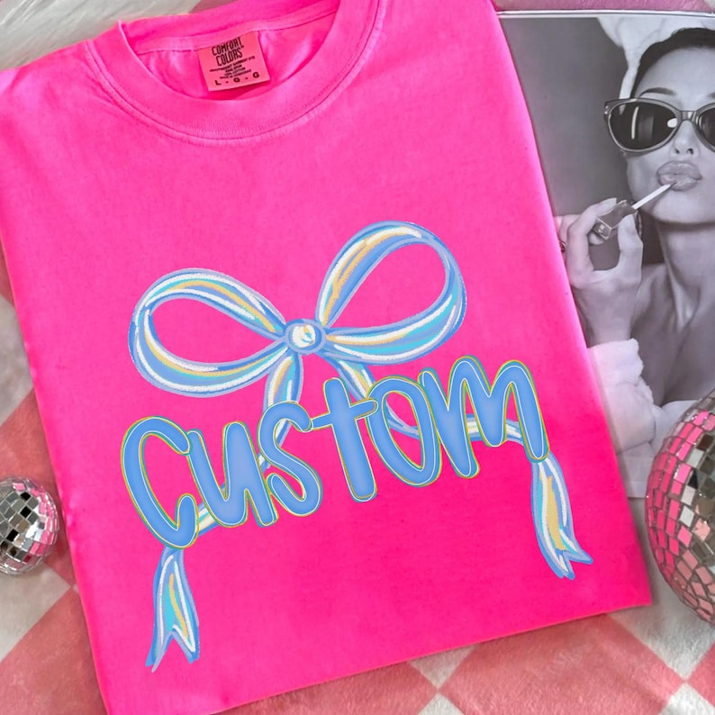 May include: A vibrant pink t-shirt featuring a blue and yellow bow graphic and the word "Custom" in blue. The shirt is displayed on a pink and white checked surface. A monochrome photo is in the background.