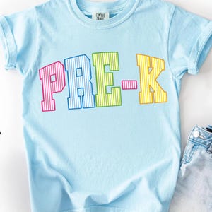 May include: A light blue short-sleeve t-shirt featuring the text PRE-K in large, block-style letters. Each letter has a vertical striped pattern in alternating colors of pink, blue, green, and yellow. The shirt has a crew neckline.