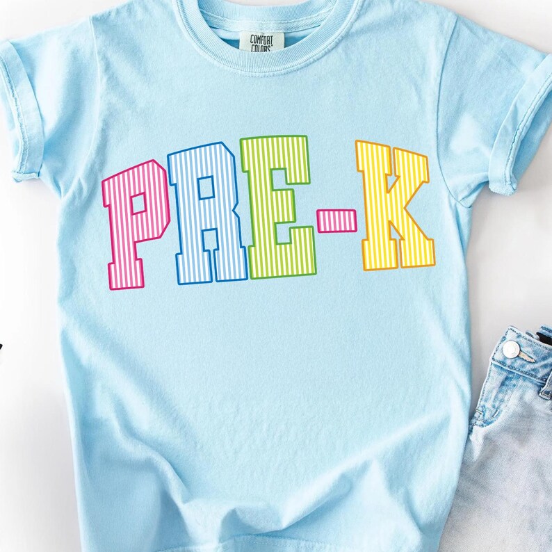 May include: A light blue short-sleeve t-shirt featuring the text PRE-K in large, block-style letters. Each letter has a vertical striped pattern in alternating colors of pink, blue, green, and yellow. The shirt has a crew neckline.
