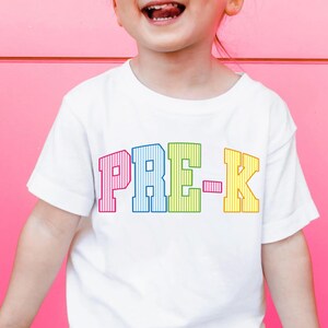 May include: A white short-sleeved t-shirt featuring the text PRE-K printed in a bold, arched collegiate font. Each letter is filled with a vertical striped pattern in distinct colors: pink for P, blue for R, green for E, and yellow for K.