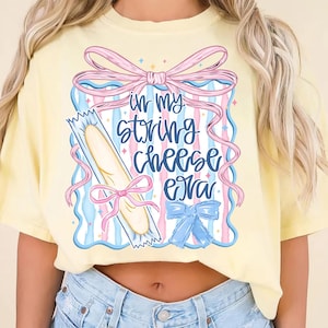 May include: A light yellow t-shirt with a graphic that says "in my string cheese era." The graphic features a string cheese stick, pink and blue bows, and a blue and pink striped background.