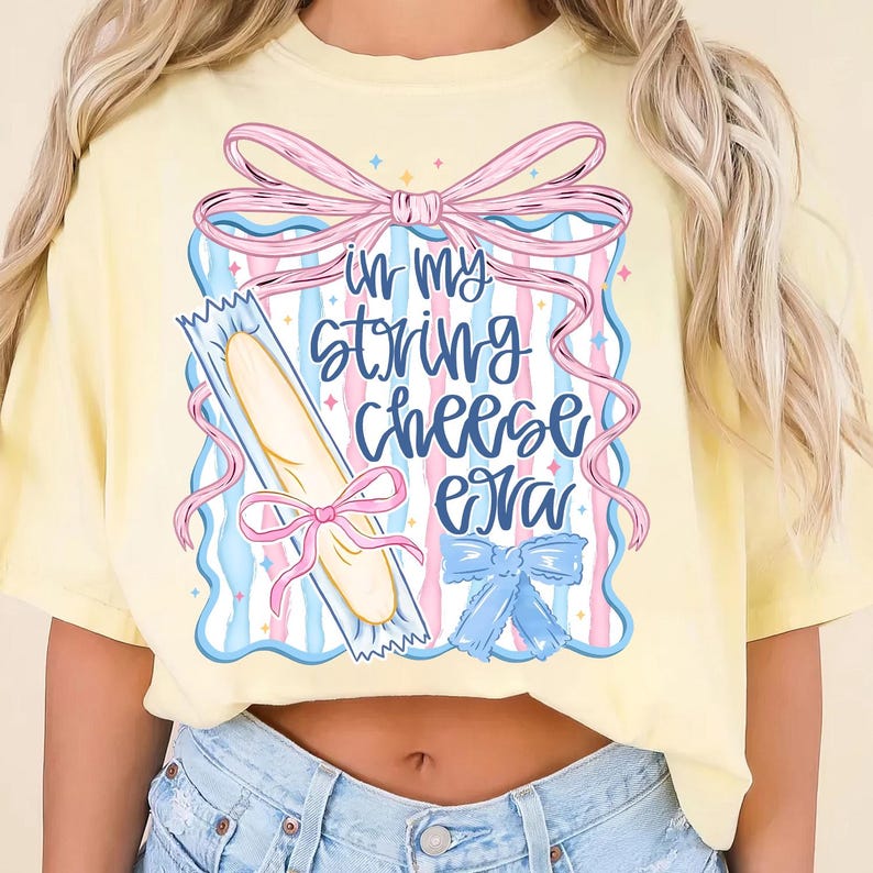 May include: A pale yellow t-shirt with the phrase "in my string cheese era" printed on it. The design includes a string cheese stick, pink and blue bows, and a blue and pink striped background.