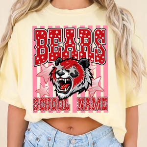 May include: A light yellow t-shirt featuring a graphic of a bear with star-shaped eyes and the word "BEARS" in red letters. The design includes pink and white stripes and the text "SCHOOL NAME".
