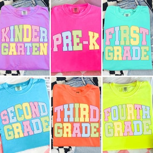 May include: A collection of six short-sleeved crew neck t-shirts in various bright colours including lavender, hot pink, teal, light blue, orange, and neon yellow. Each shirt features block lettering with vertical stripes in pastel colours.
